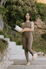 Woman in brown athletic wear holding a yoga mat on stone steps with greenery in the background