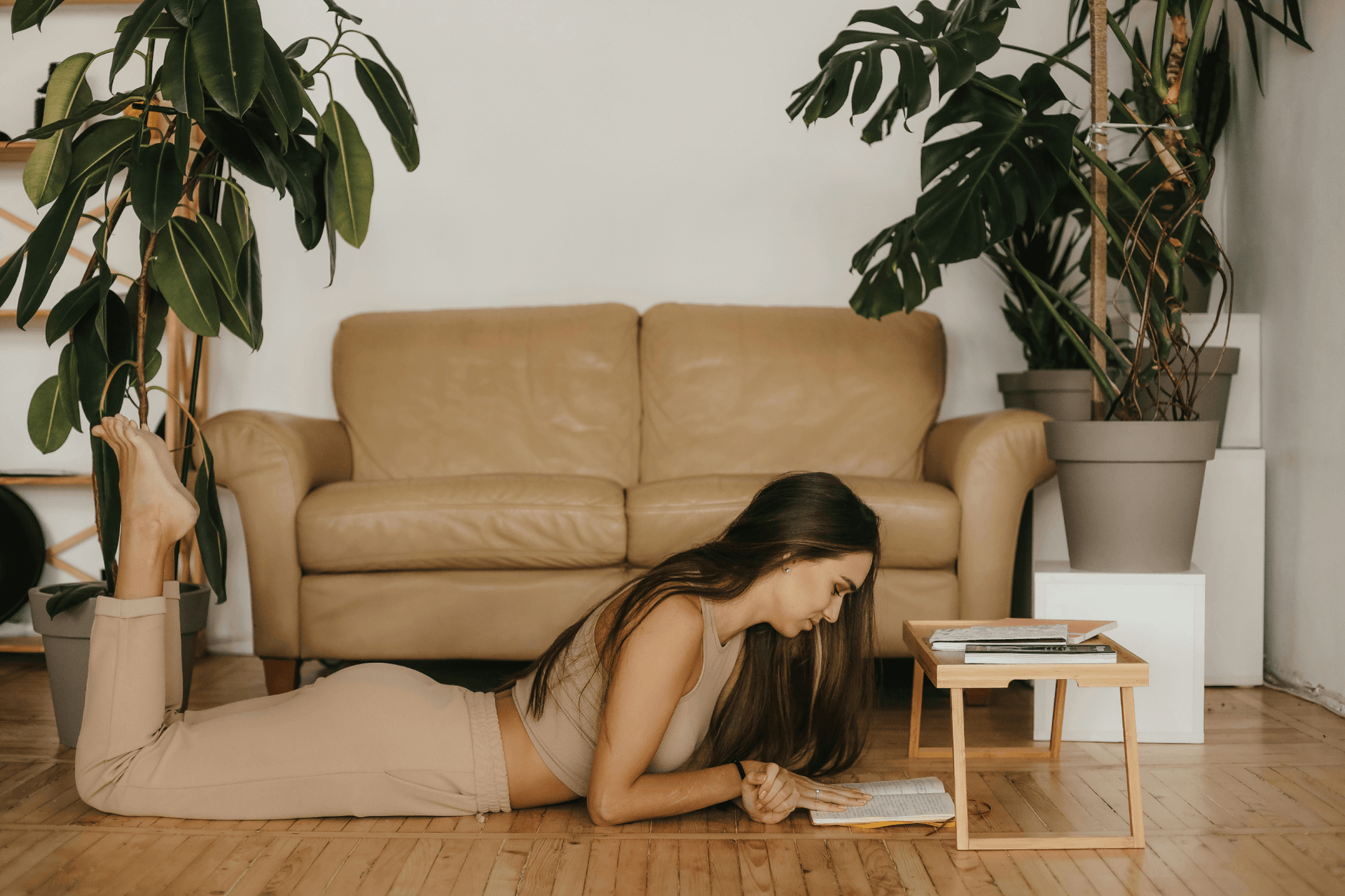 Woman lying on the floor reading a book in a cozy living room with plants and a couch.