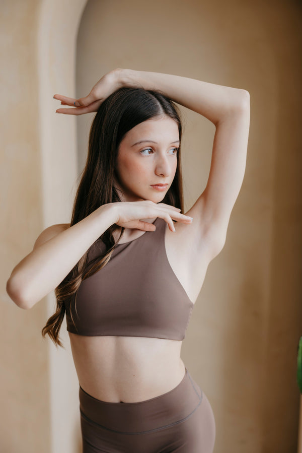 Woman in a brown sports bra and leggings posing against a beige wall.